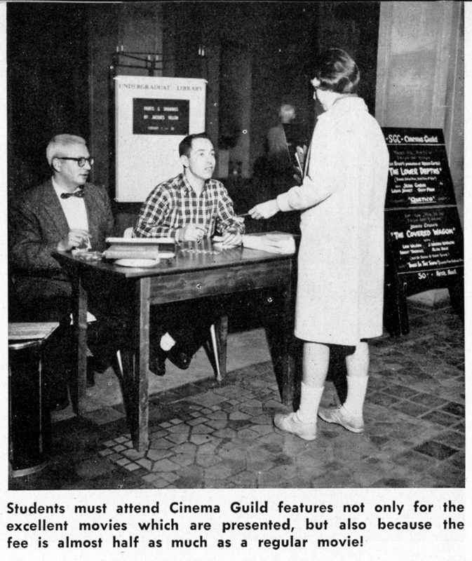 An old photo of two men sitting at a table selling tickets, and a woman standing in front of the table buying a ticket.