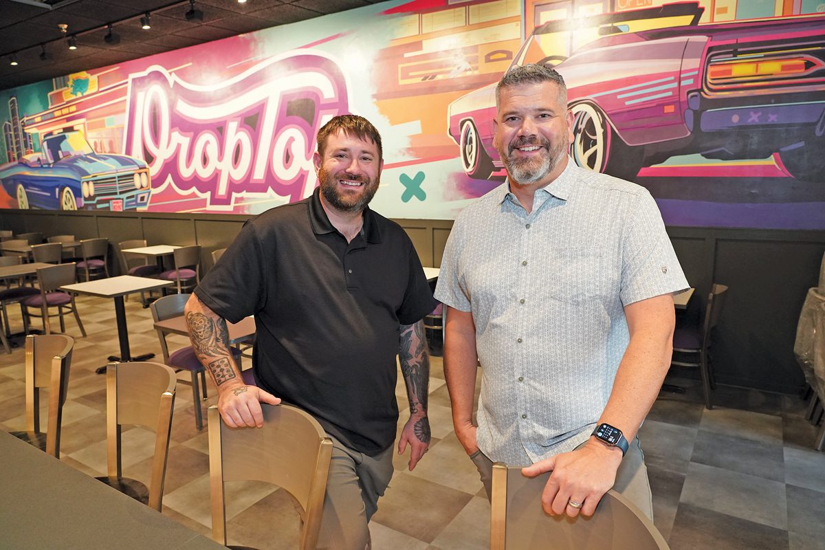 Two men standing in a bar with the word DropTop mural on the wall.