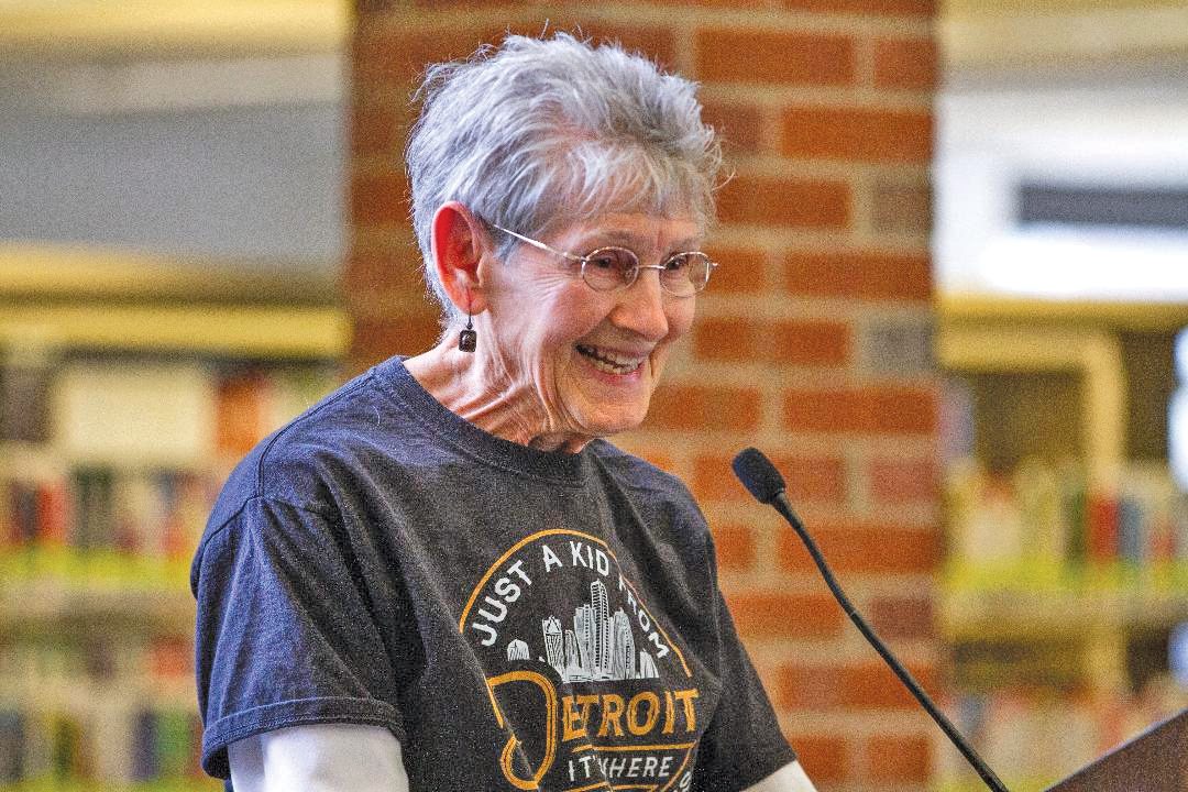 Pauline Loewenhardt smiling and standing at a microphone. She has short gray hair and is wearing glasses and a gray t-shirt.