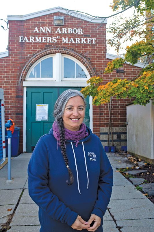 Stefanie Stauffer smiling in front of the Ann Arbor Farmers' Market building. She is wearing a winter jacket and her hair is in a braid.