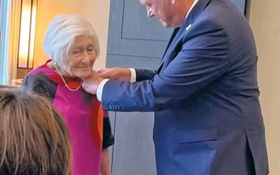 A tall bald man in a suit is pinning a medal to the lapel of a short woman in a magenta top with white hair.