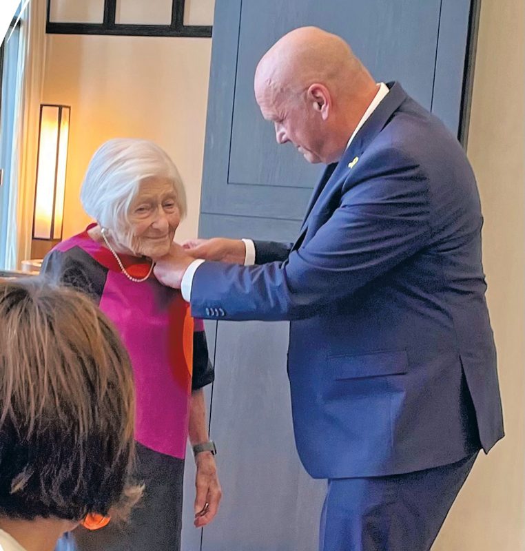A tall bald man in a suit is pinning a medal to the lapel of a short woman in a magenta top with white hair.