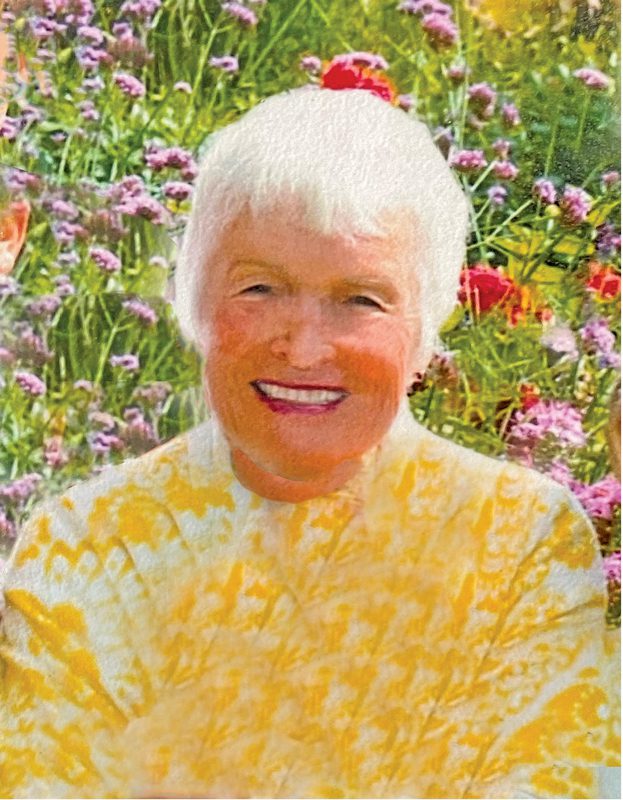 An older woman standing in a flower garden and smiling. The sun is illuminating her hair. She is wearing a yellow shirt.