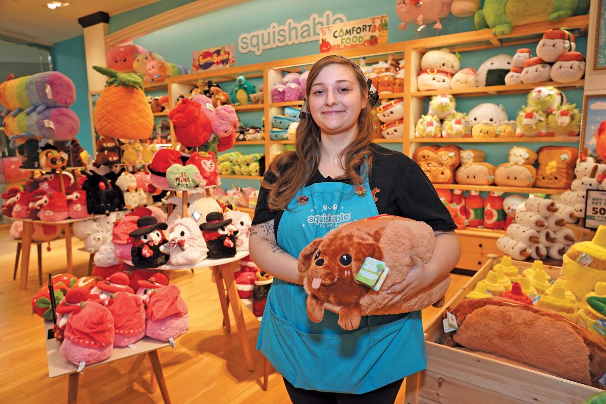 A young woman in a lue apron in a store filled with big, squishy, soft-looking stuffed animals. She's holding a stuffed dahschund the size of a pillow.