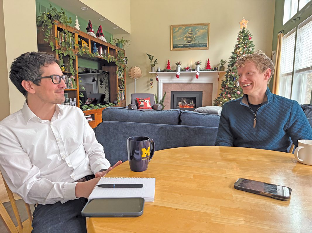 Two young men sit at a round table in a living room with Christmas decorations in the background.
