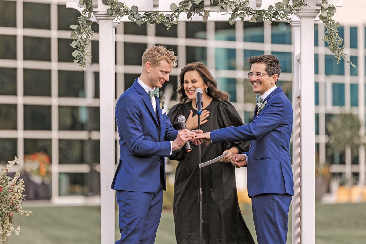 A woman is officiating a wedding between two men. The grooms are wearing blue suits, and the one on the left is putting the ring on the other's finger. The woman holds her hand to her heart. All three are smiling with joy.