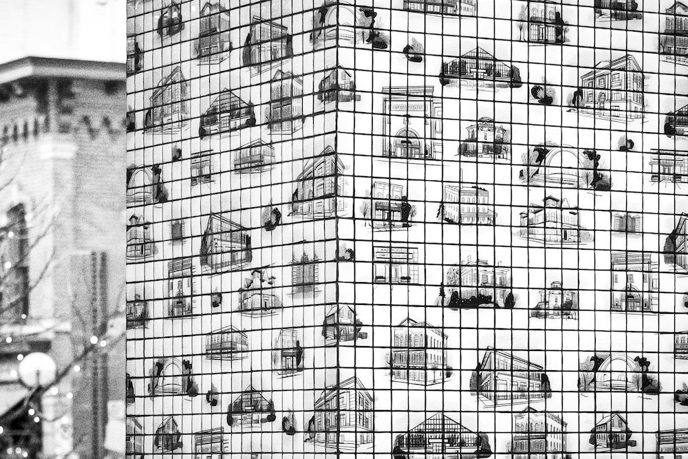 A black-and-white photo of a building with the corner in the foreground. The surface of the building is tiled, with illustrations of buildings on the tiles.