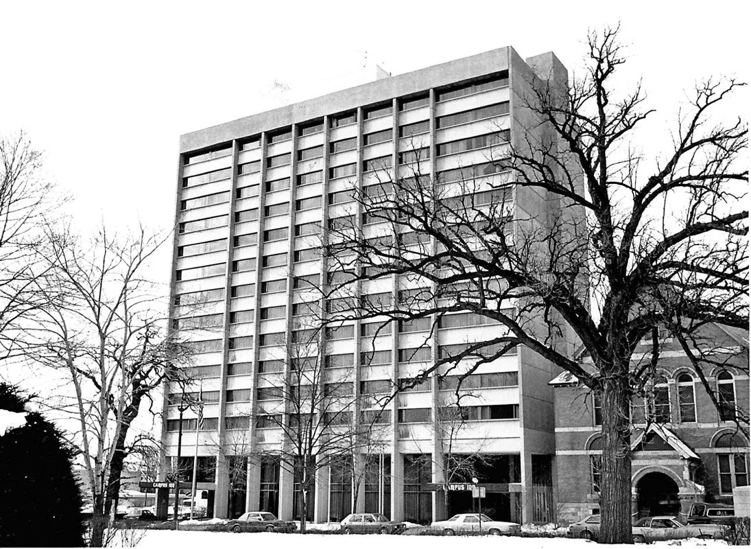 Black-and-white photo of the Campus In, a tall building of more than 10 floors, with winter trees in the foreground.