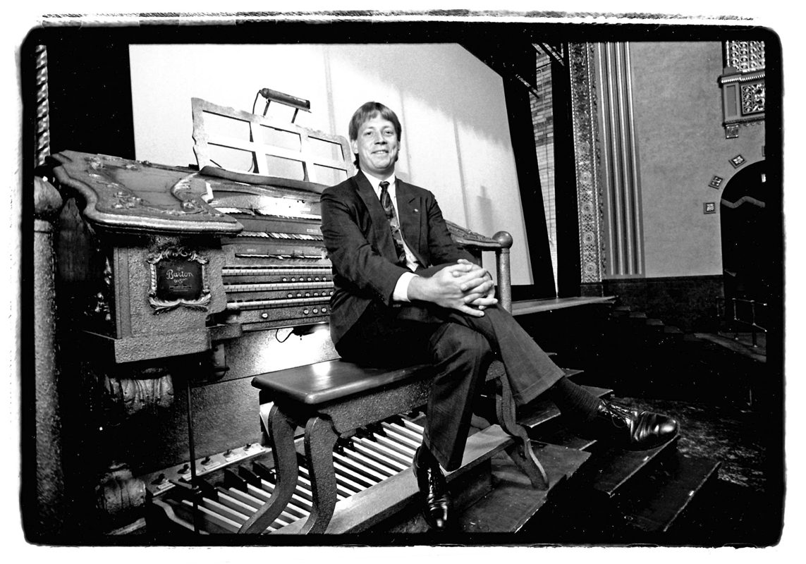 A black-and-white photo of a man sitting on an organ bench facing away from the organ.