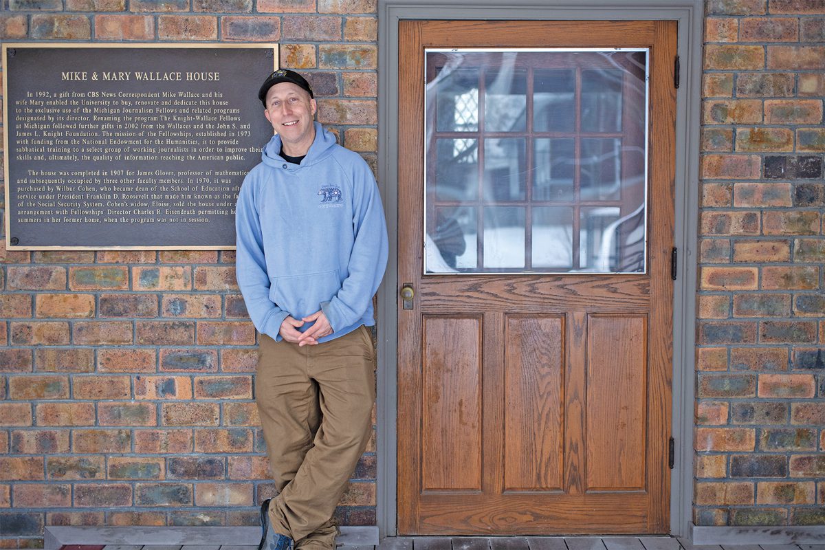 A man in a blue hoodie and a baseball cap standing in front of a brick building with a plaque and a wooden door with a window.