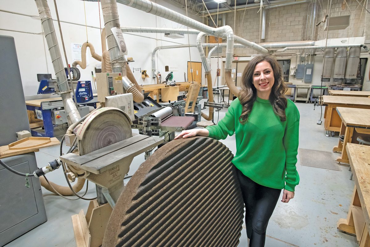 A young woman standing next to a round piece of wood with a grooved surface. She's in a large warehouse space filled with tools.