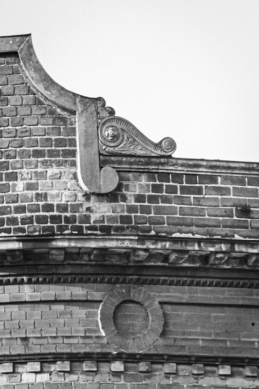 Black-and-white photo of a brick exterior of a building with a filigreed bit of decoration at the top.