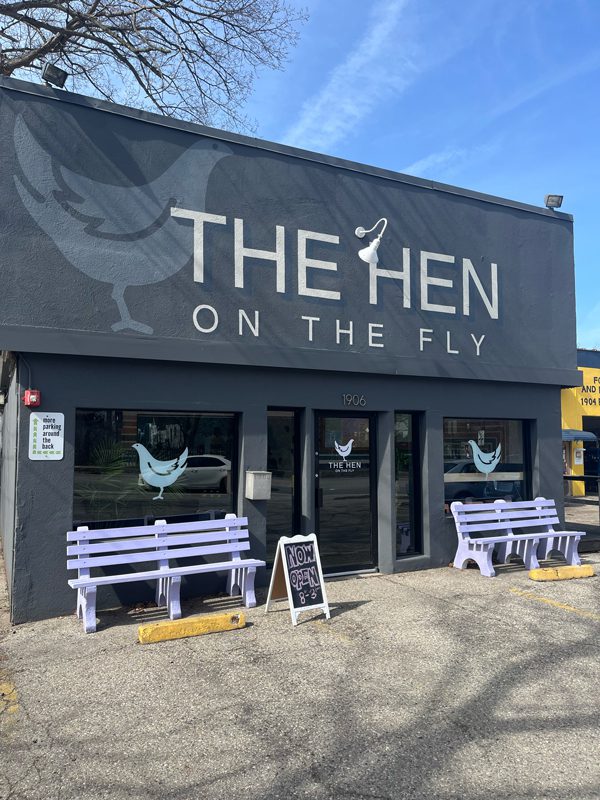 Exterior photo of The Hen on the Fly. A gray building with big windows and two purple benches out front. 