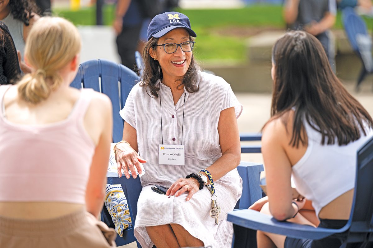 A woman in a University of Michigan baseball cap and a white dress smiles and chats with two college students whose backs are to the camera. They are sitting outside.