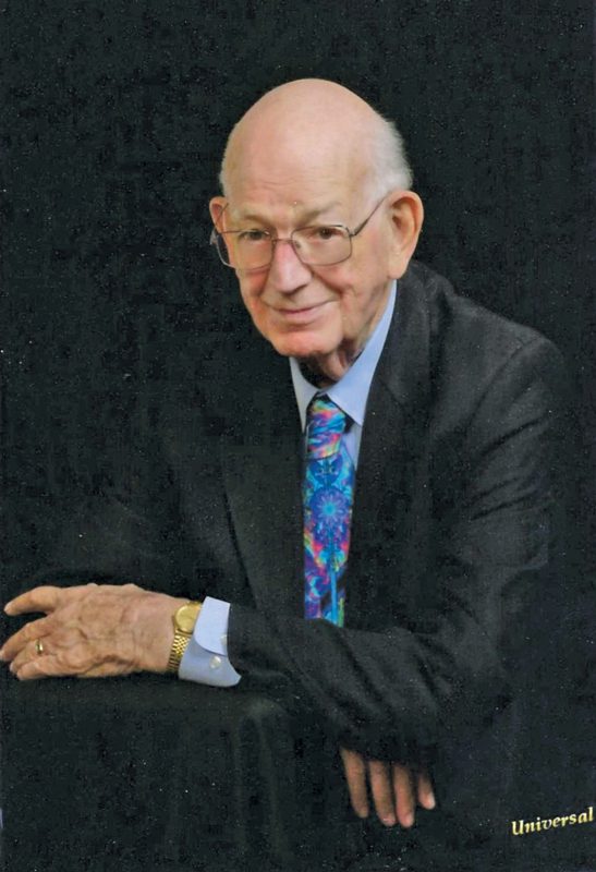 A portrait of a distinguished-looking older man wearing a suit with a colorful tie and glasses.