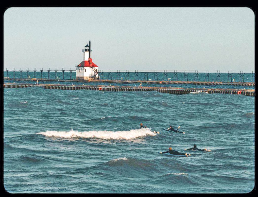An artsy photo of four surfers swimming with a lighthouse in the background