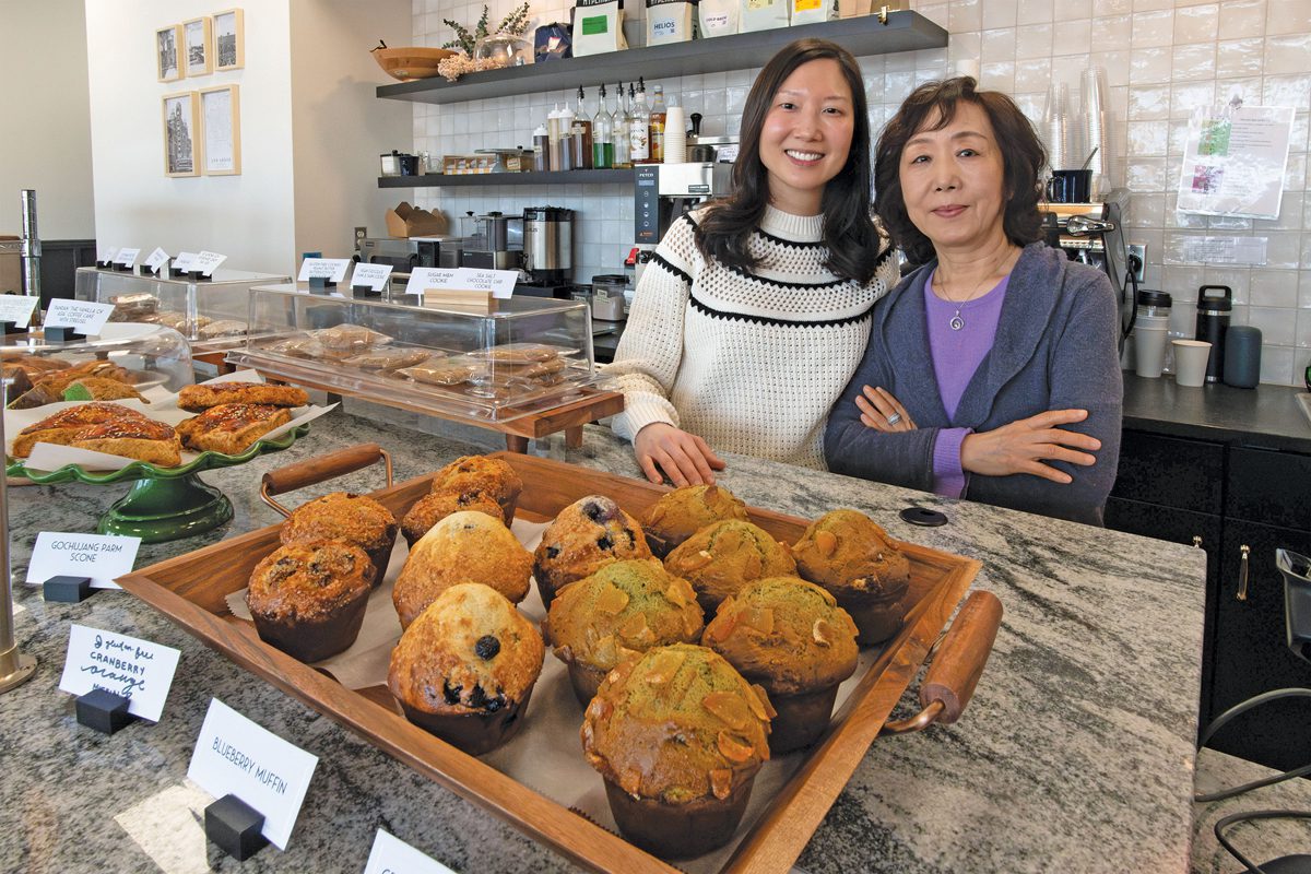 Two women standing in a bakery with a tray of muffins in the foreground