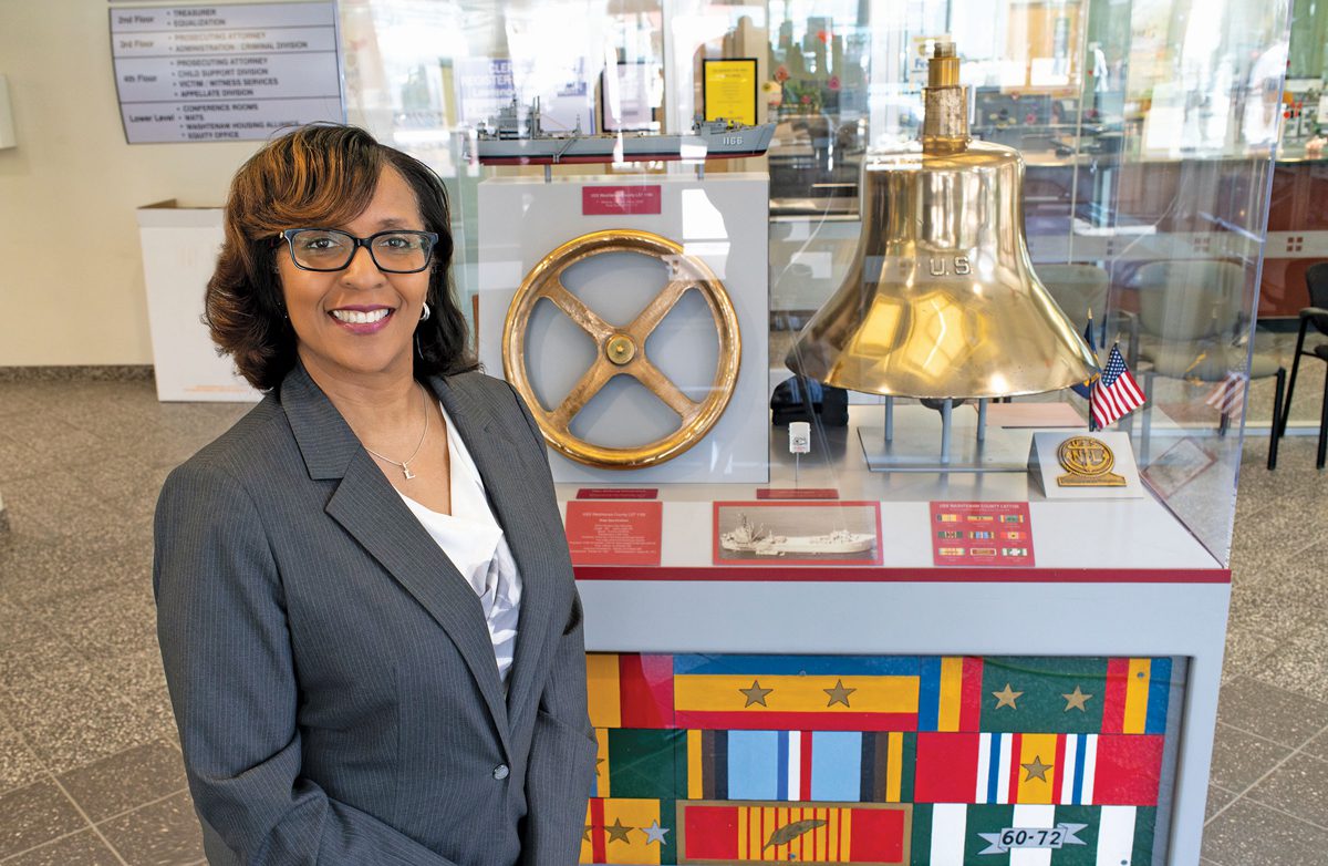 A woman in a suit jacket and glasses smiling in front of a bell.