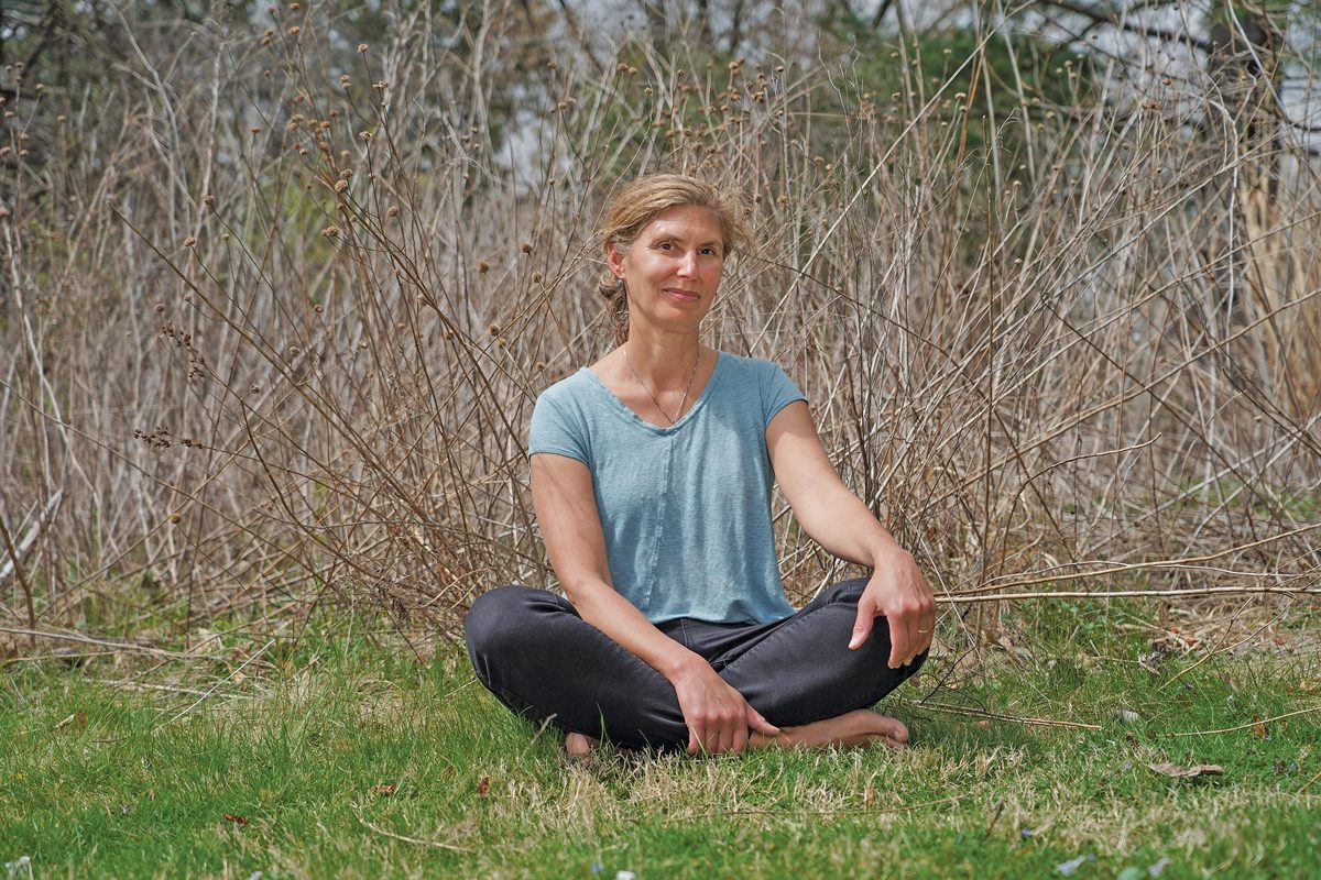 A barefoot woman sitting cross-legged in a field with a wise smile on her face.