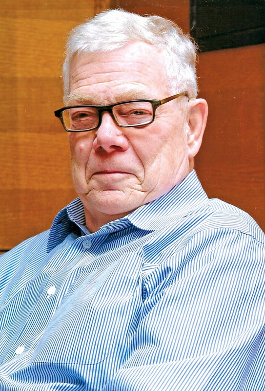 A distinguished-looking older man with glasses and white hair, wearing a blue-and-white striped collared shirt. 