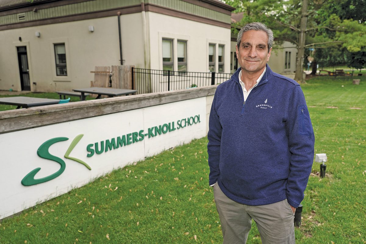 The head of Greenhills School standing in front of a sign for Summers-Knoll school. He has his hands in his pockets and a somber look on his face.