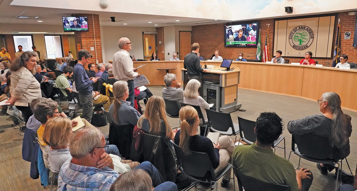 A meeting in a government building. People sit in rows in chairs facing a group of administrators at a desk. There is a podium, and people are lined up to speak. 