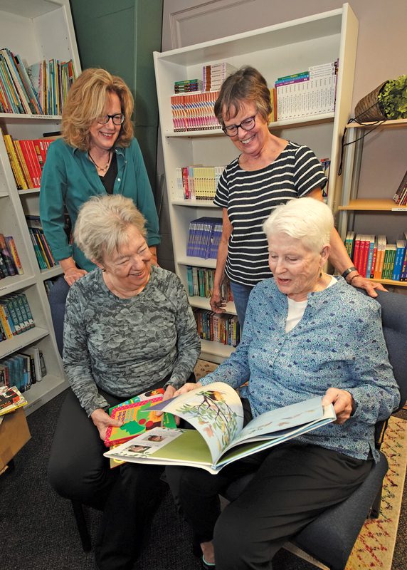 Four women in a library. Two of them are standing in the background, and two are sitting in the foreground reading a children's book.