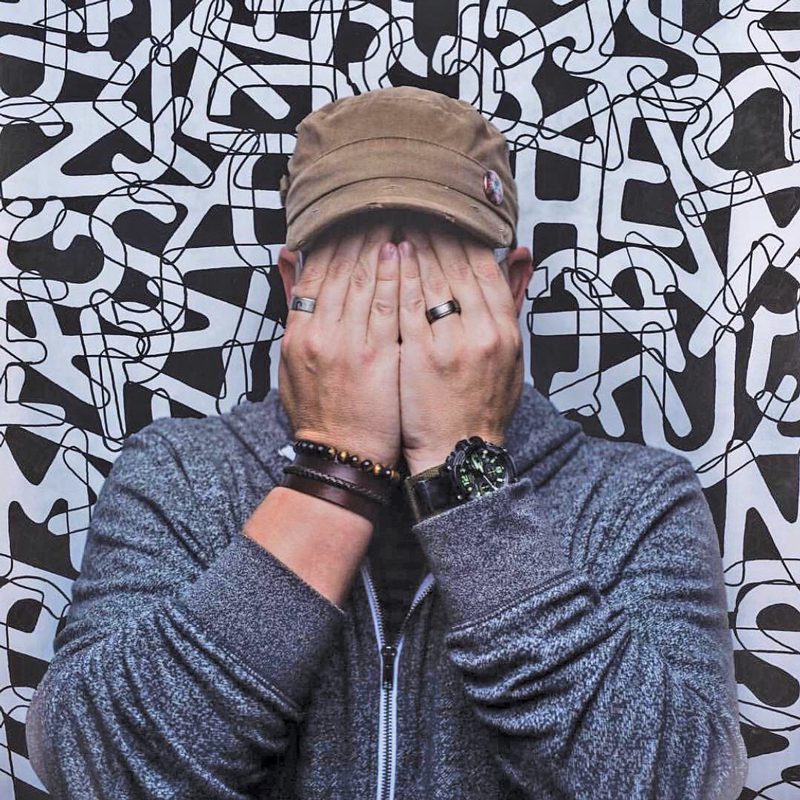 Photograph of a man from the chest up. He's covering his face iwth his hands. In the background are overlapping letters. 