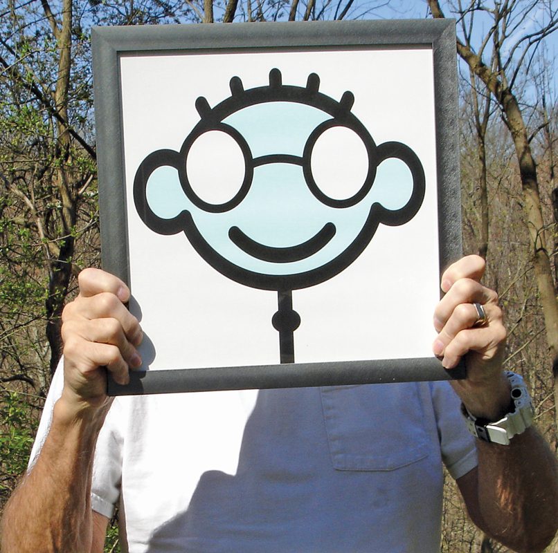 Photo of a man outside. He's holding up a framed cartoon of a smiling face with glasses over his own face.