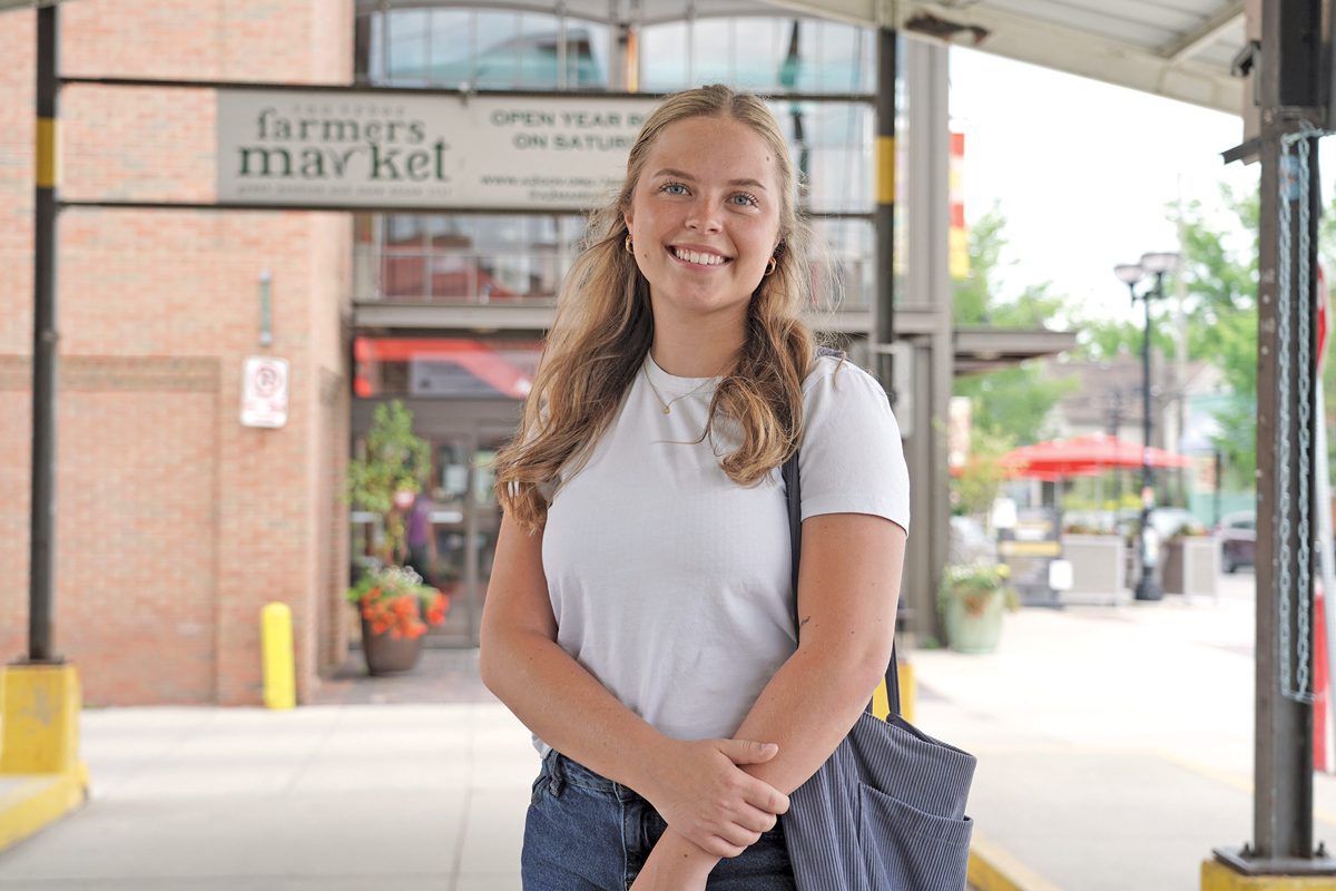 A girl standing in front of a farmer's market. She's smiling and wearing a white T-shirt. Her arms are crossed in front of her.
