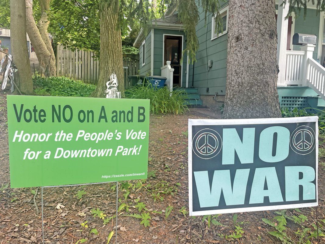 Signs in a yard. one says Vote NO on A and B: Honor the People's Vote for a Downtown Park. The other says NO WAR.