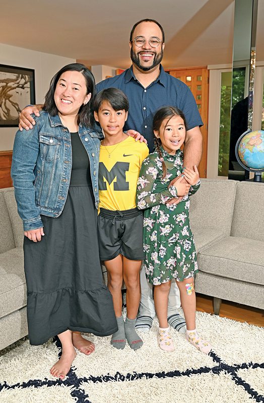 A family standing in their living room. The mother is on the left, wearing a long dress and a jean jacket. The father has his arms around her and the two kids.