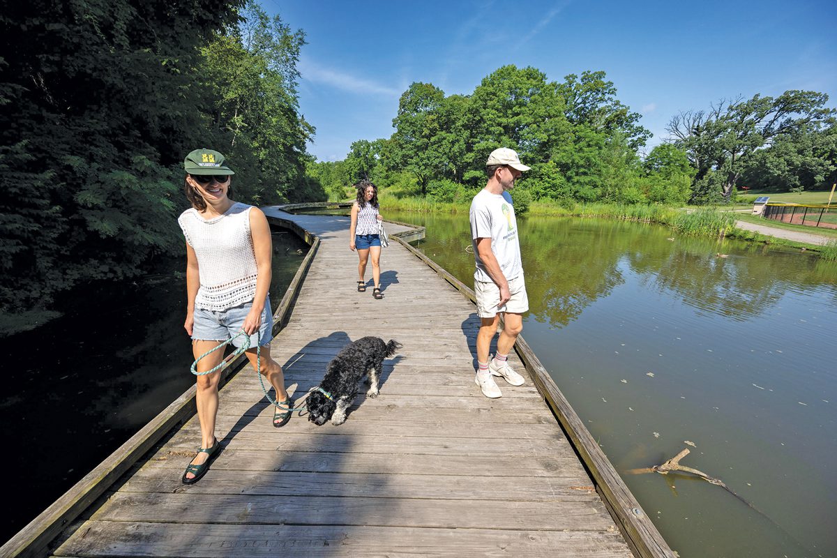 Three people and a dog standing on a boardwalk over a pond.