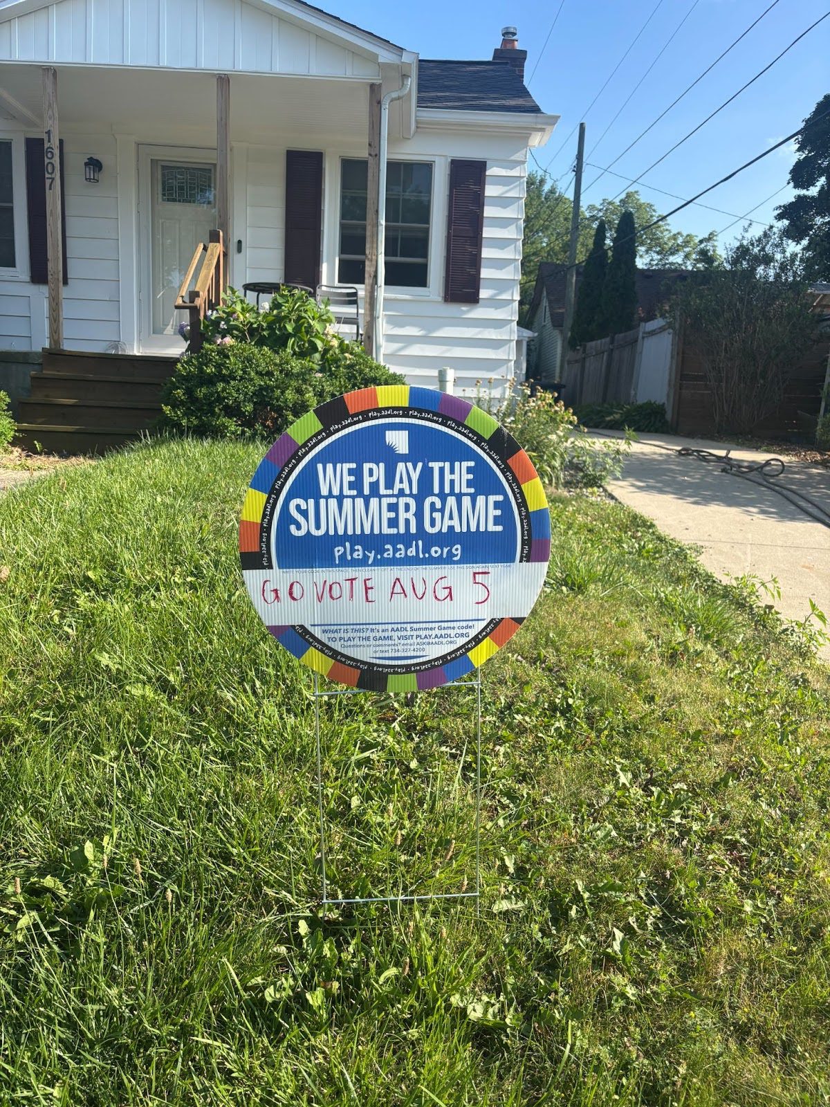 A circular sign in the front yard of a white house. It reads: We play the summer game: Go vote Aug. 5.