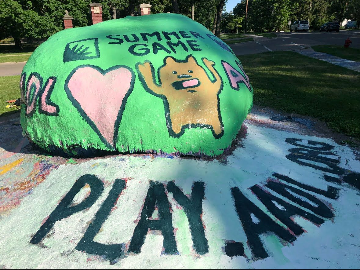 A boulder painted green with a heart, a bear, the AADL logo, and the words "Summer Game." On the sidewalk, it says play.aadl.org.