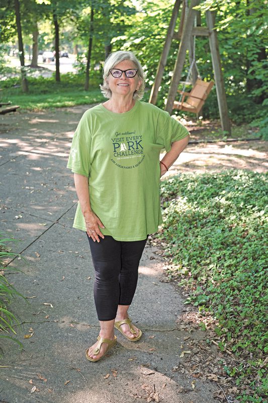 A smiling woman with gray hair and glasses stands in a park with a swinging bench. 