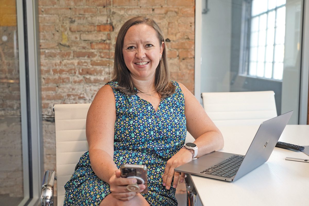 a woman smiles at a laptop at a desk, holding a cellphone in her hand