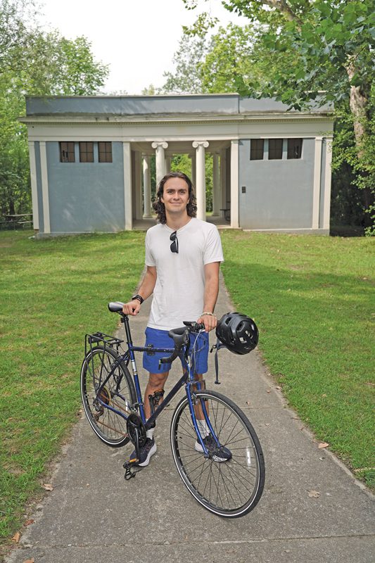 A young man stands with a bicycle in a park with a building that has Greek columns. 