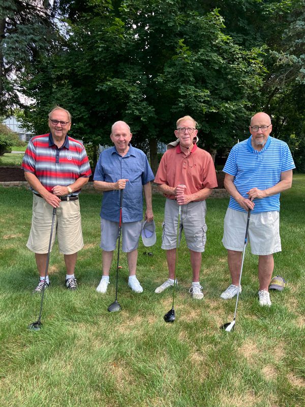 Four older men standing in a golf course holding golf clubs.