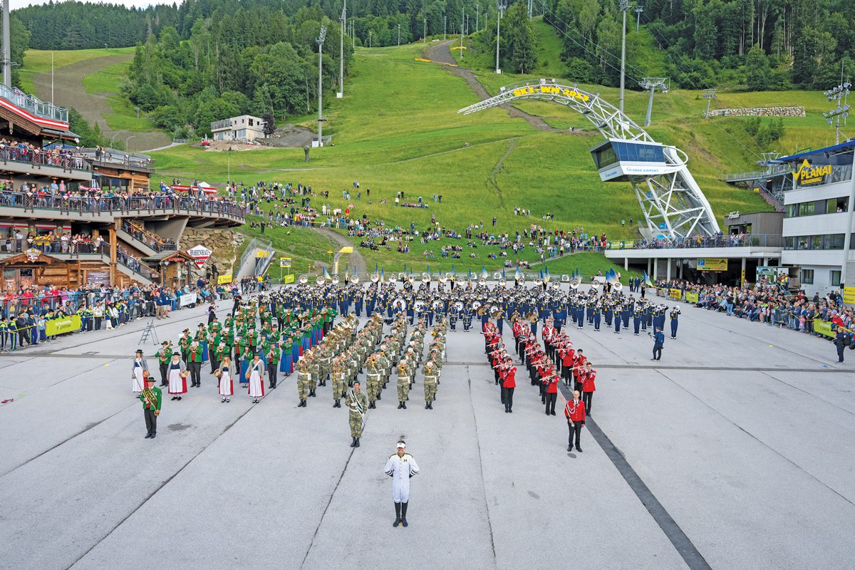 Three marching bands in a public square with a hill in the background