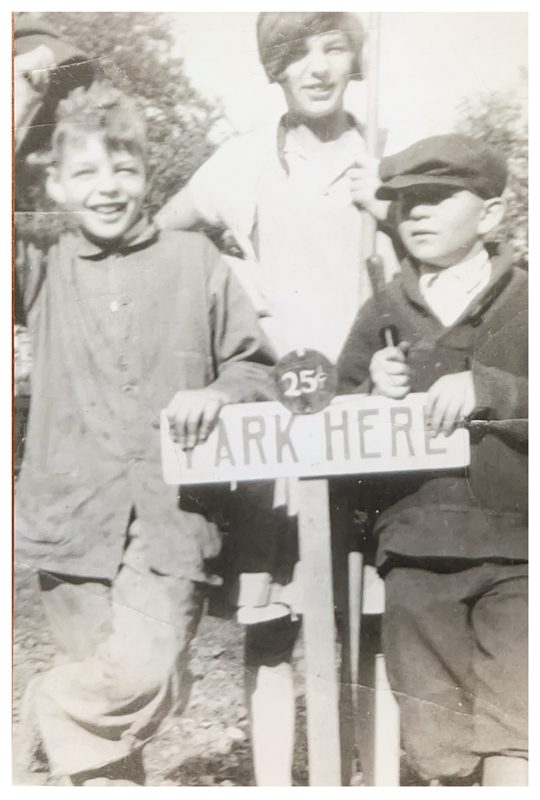 Black-and-white photo of three children in old-fashioned clothing behind a sign saying "Park Here"