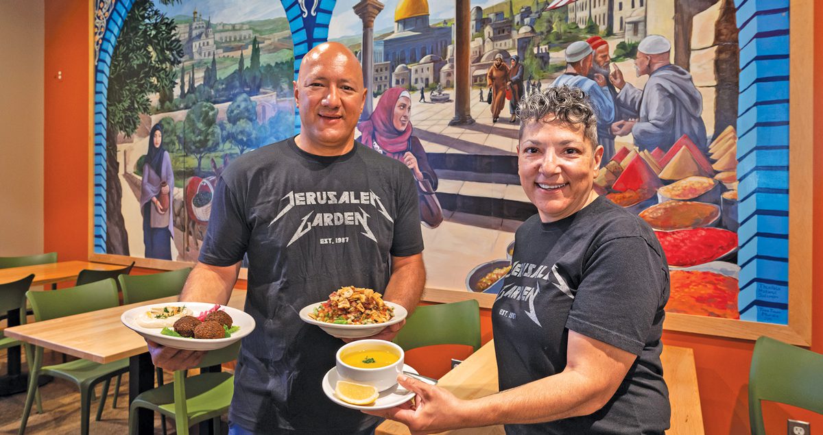 A man and woman stand in a brightly colored restaurant. They're holding an assortment of Middle Eastern food. 