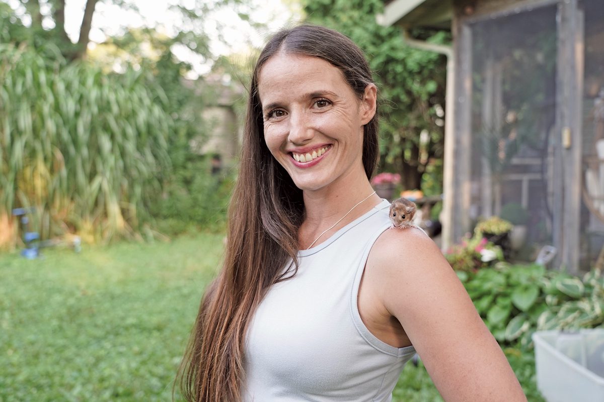 A smiling woman stands in a green yard with a mouse perched on her shoulder