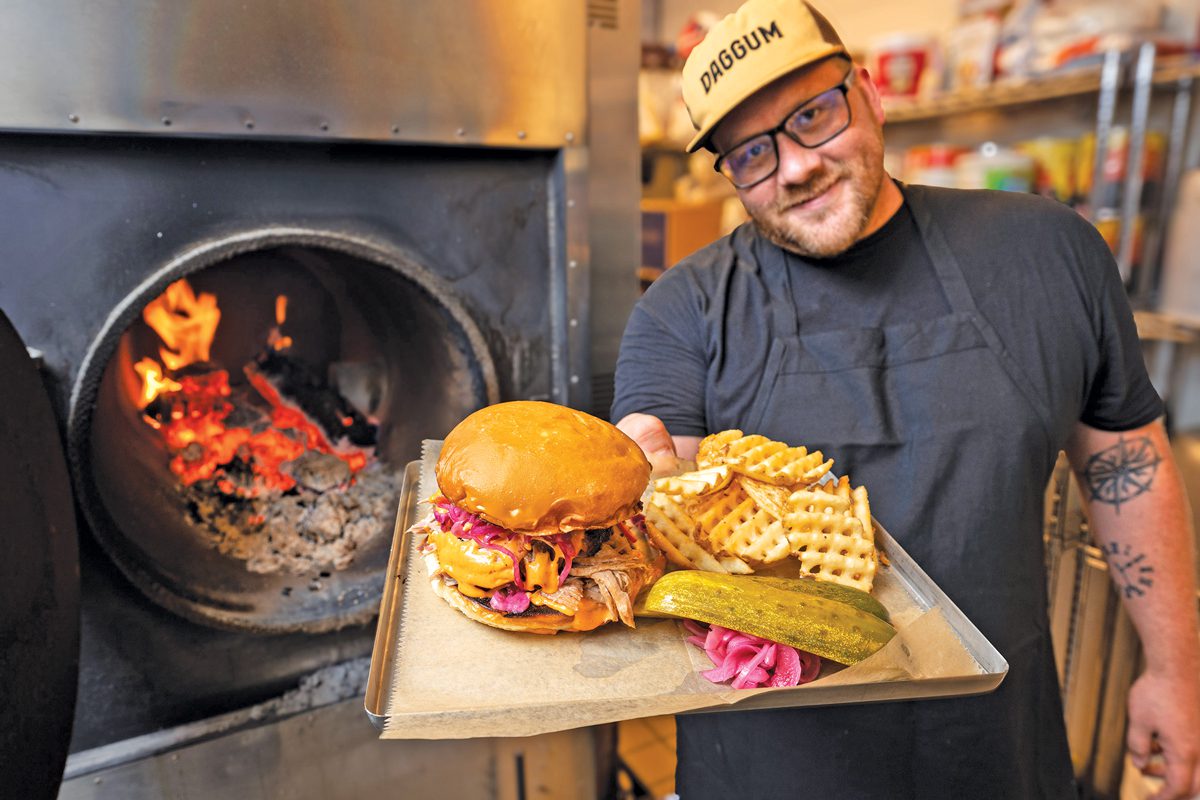 A man stands in front of a wood-fire oven. He's holding a platter with a sandwich, fries, and a pickle.