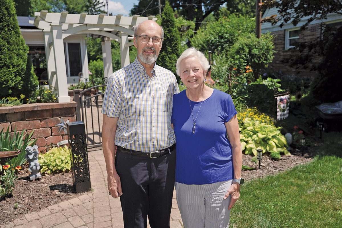 An older man and woman stand in front of a beautiful backyard garden. 