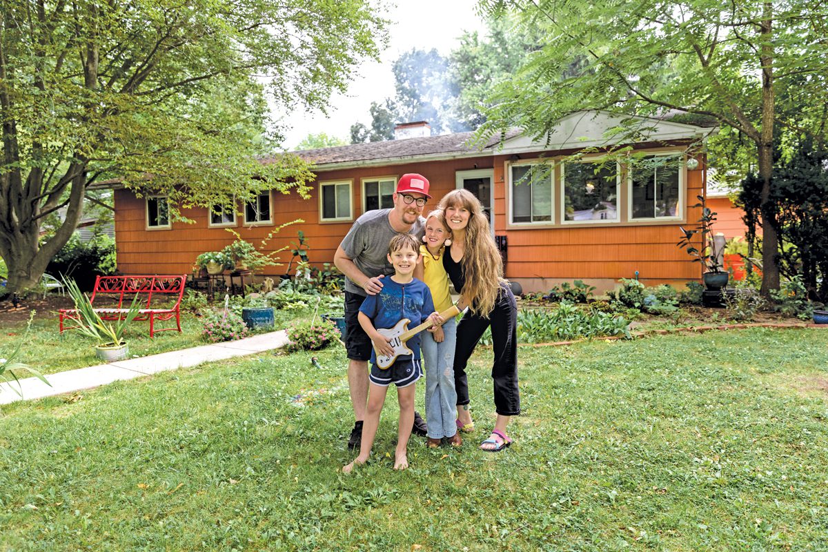 A man in a red baseball cap, a woman with long hair, a girl, and a boy holding a wooden guitar stand in the yard of an orange house