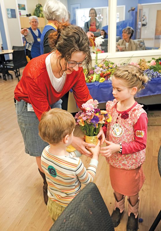 A woman bends over to hand a canister of flowers to two children.