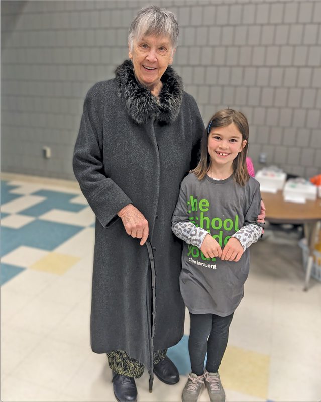 A gray-haired woman poses with a child in a school hallway.