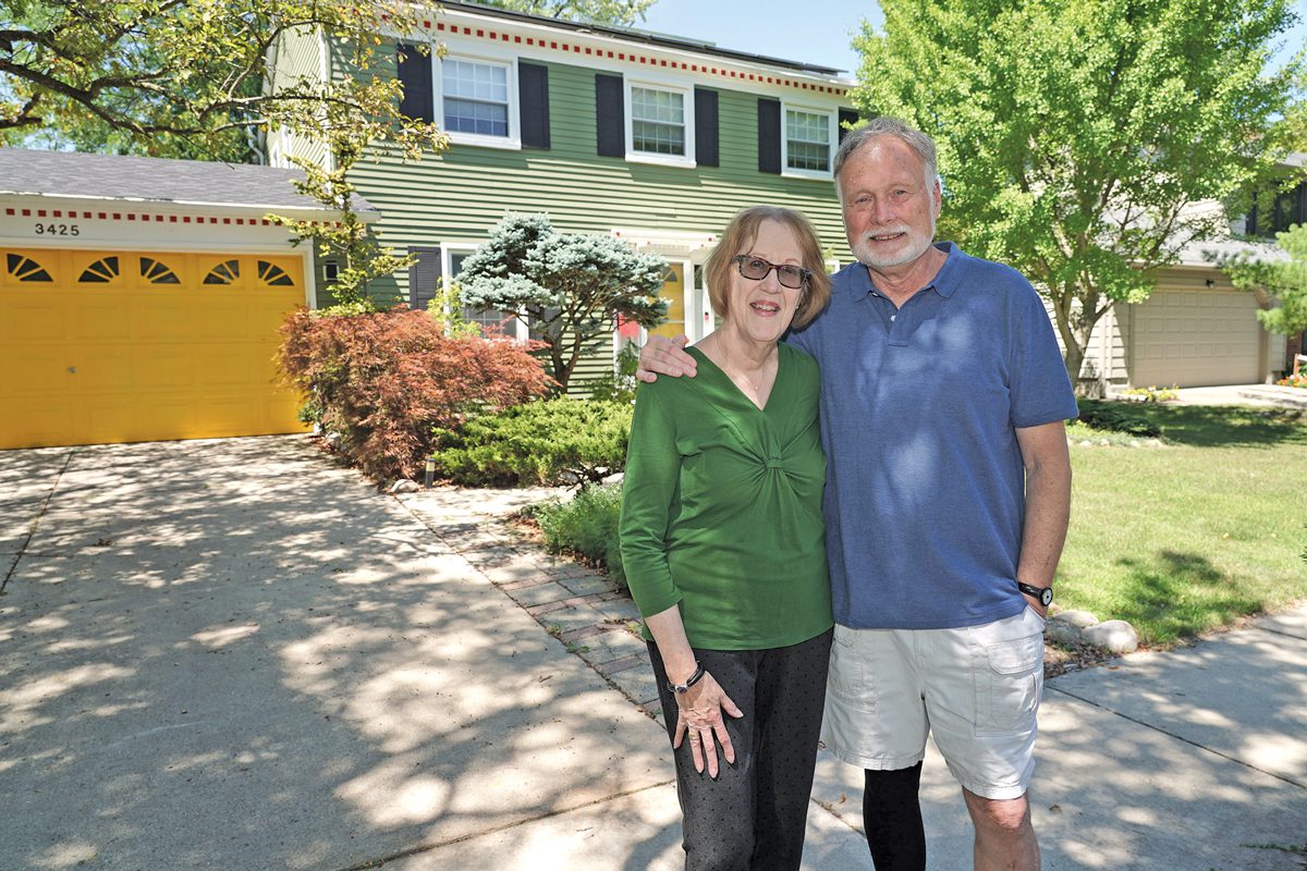 An older man and woman stand in front of a yellow house. 