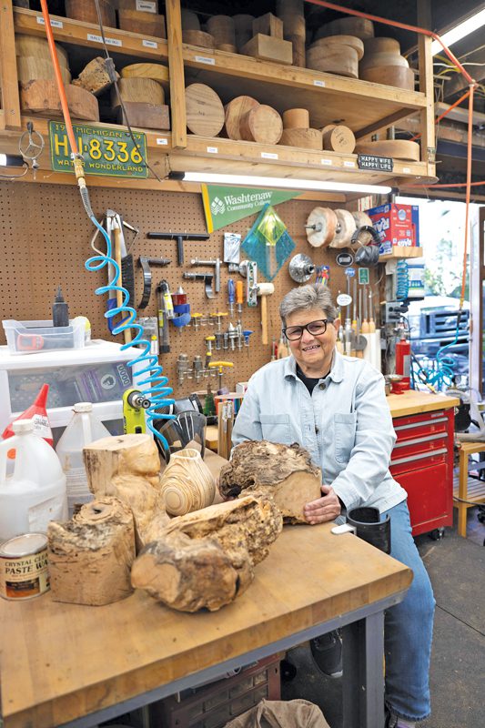 Susan Kizer sitting in a woodshop, surrounded by tools and wood of many shapes and sizes